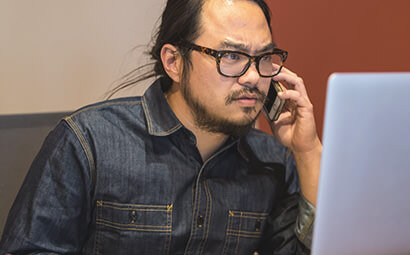Man wearing glasses and denim, sitting at a computer while talking on the phone and thinking.